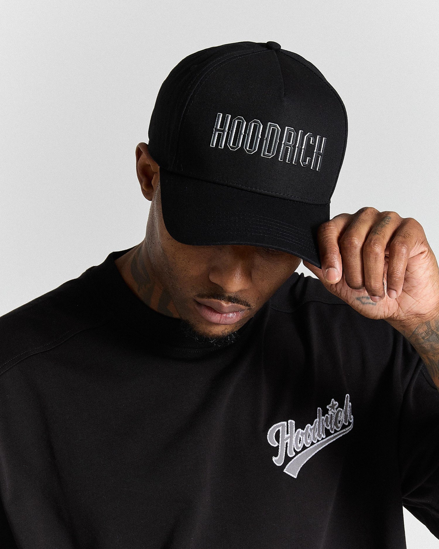 Person wearing a black cap and black hoodie with 'Hoodrich' branding on a light gray background