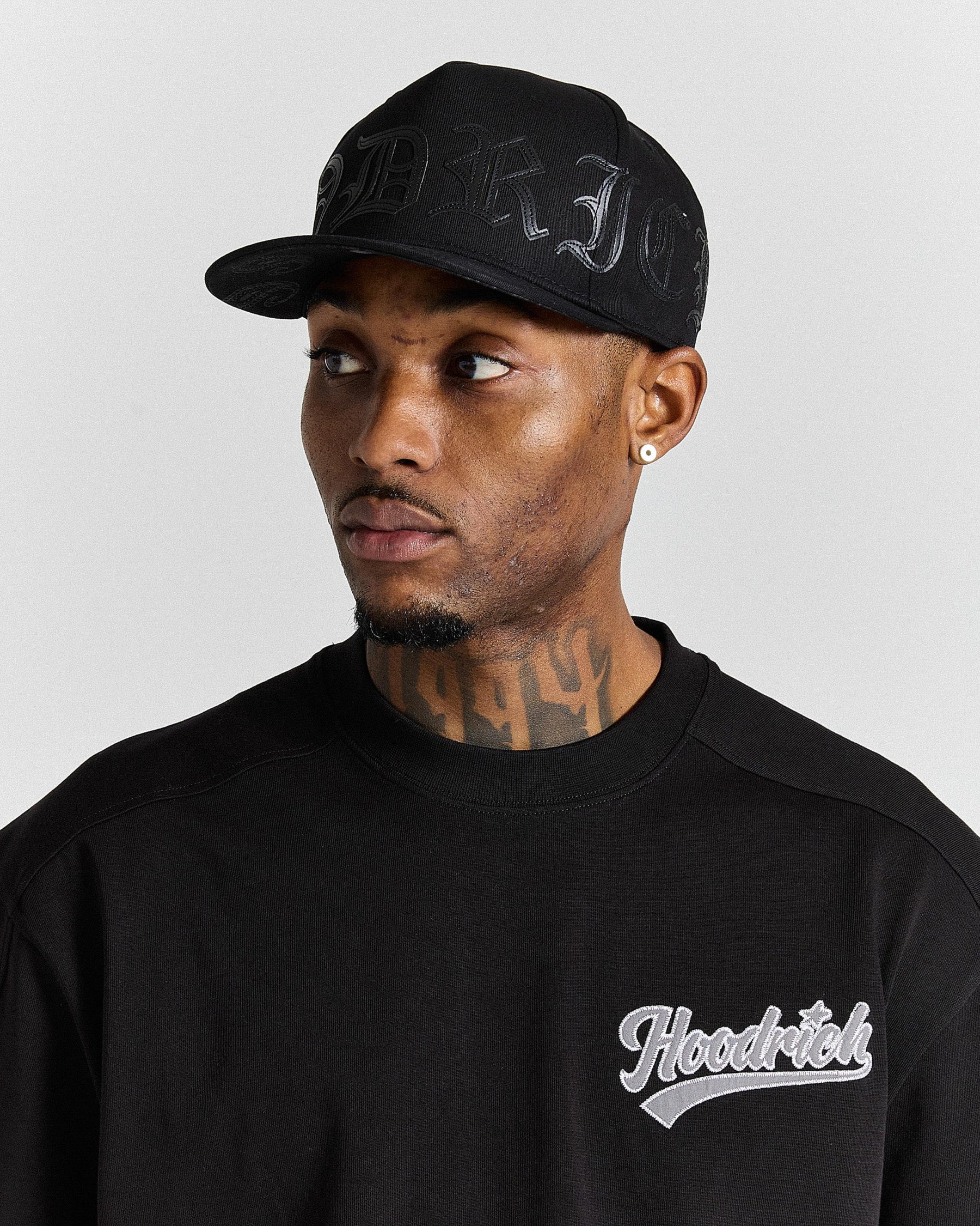 Man wearing a black cap and black 'Hoodrich' sweatshirt on a light gray background