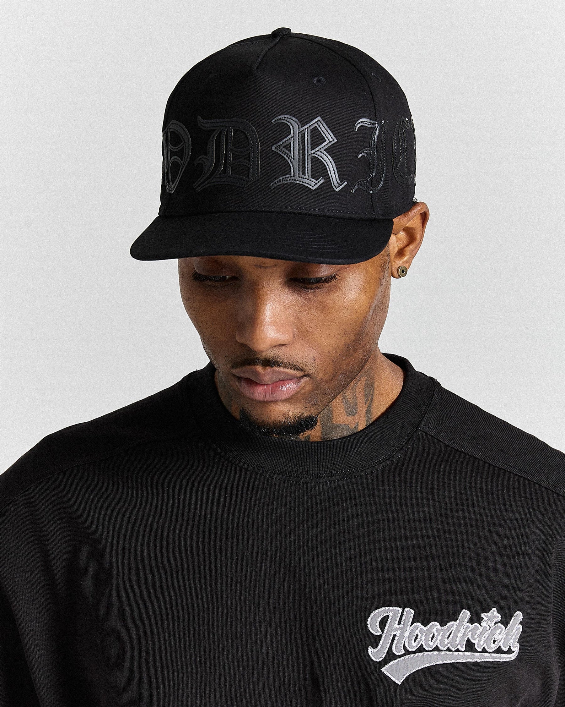 Man wearing a black cap with a black hoodie with 'Hoodrich' logo on gray background