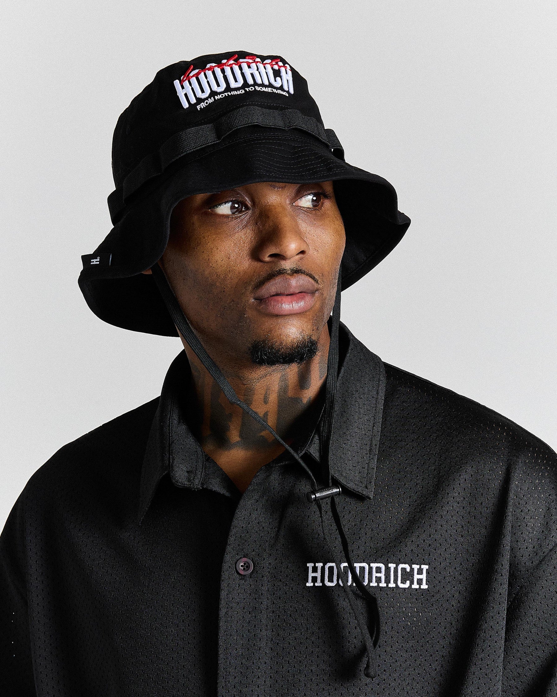 Person wearing a black 'Hoodrich' bucket hat and shirt against a gray background