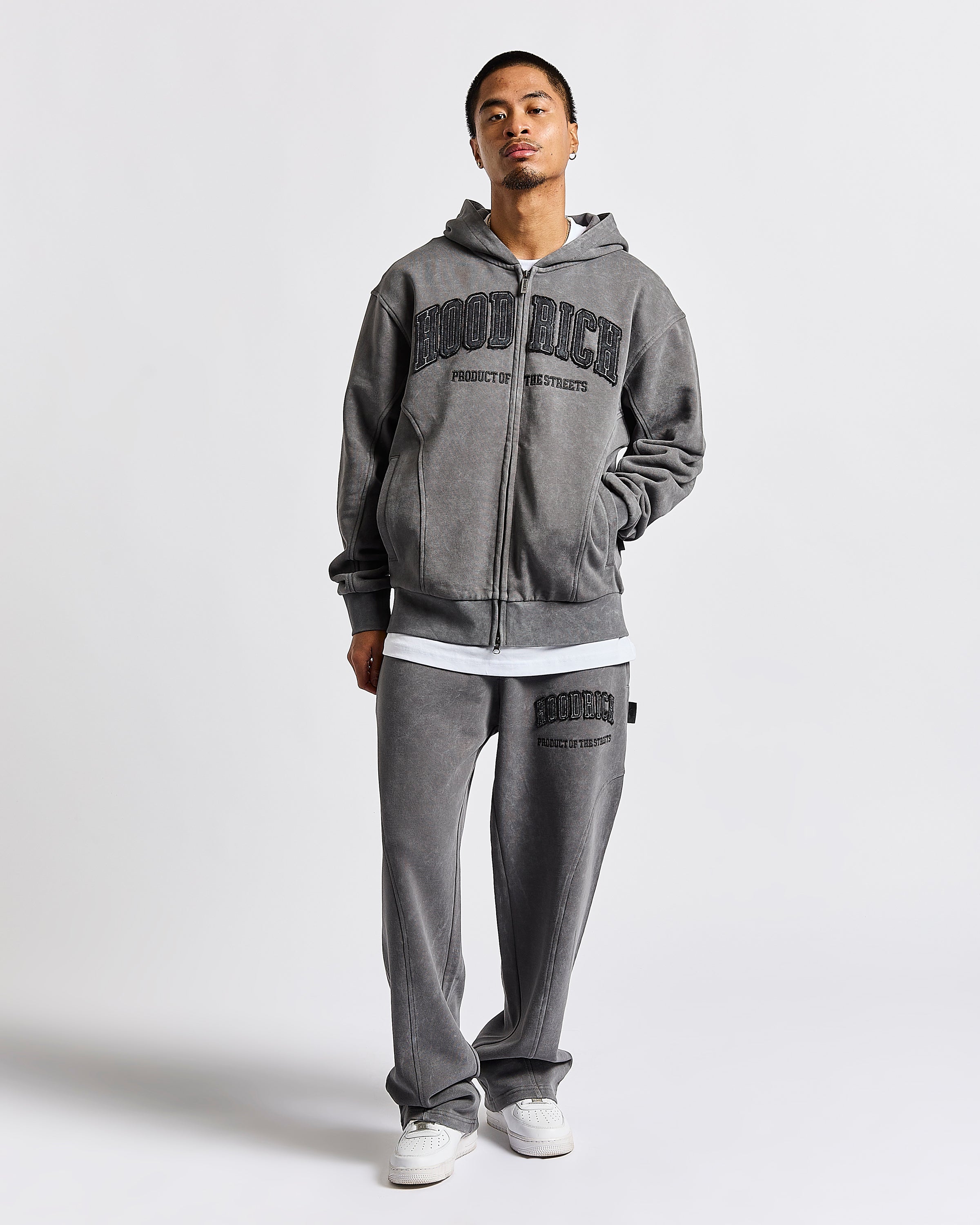 A man modeling a gray zip-up hoodie and matching sweatpants set from Good Rich, featuring black collegiate-style lettering on the front and thigh, styled with white sneakers against a light gray studio background.
