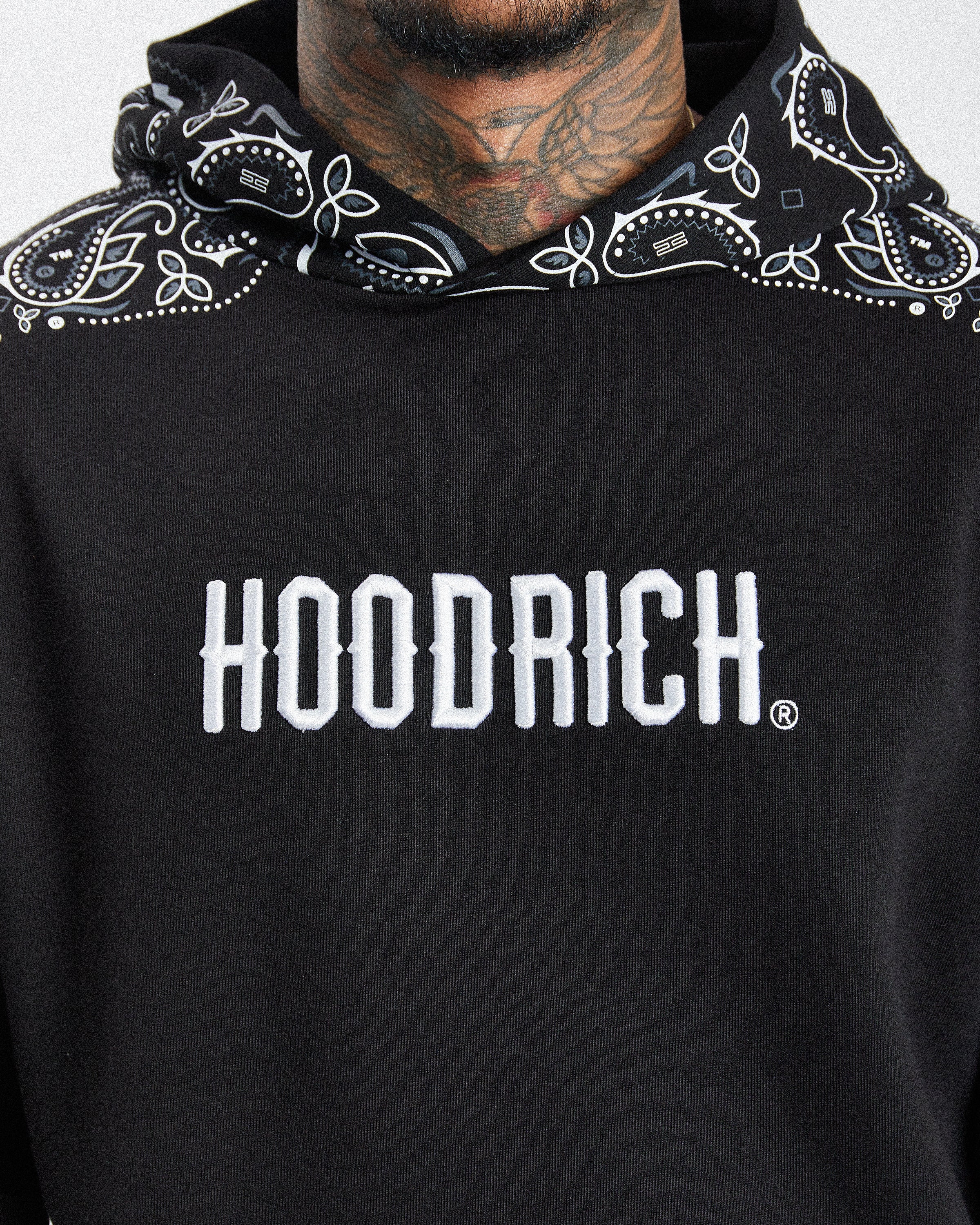 A close-up of a person wearing a black hoodie with a white 'HOODRICH' logo across the chest and a black bandana patterned hood lining.