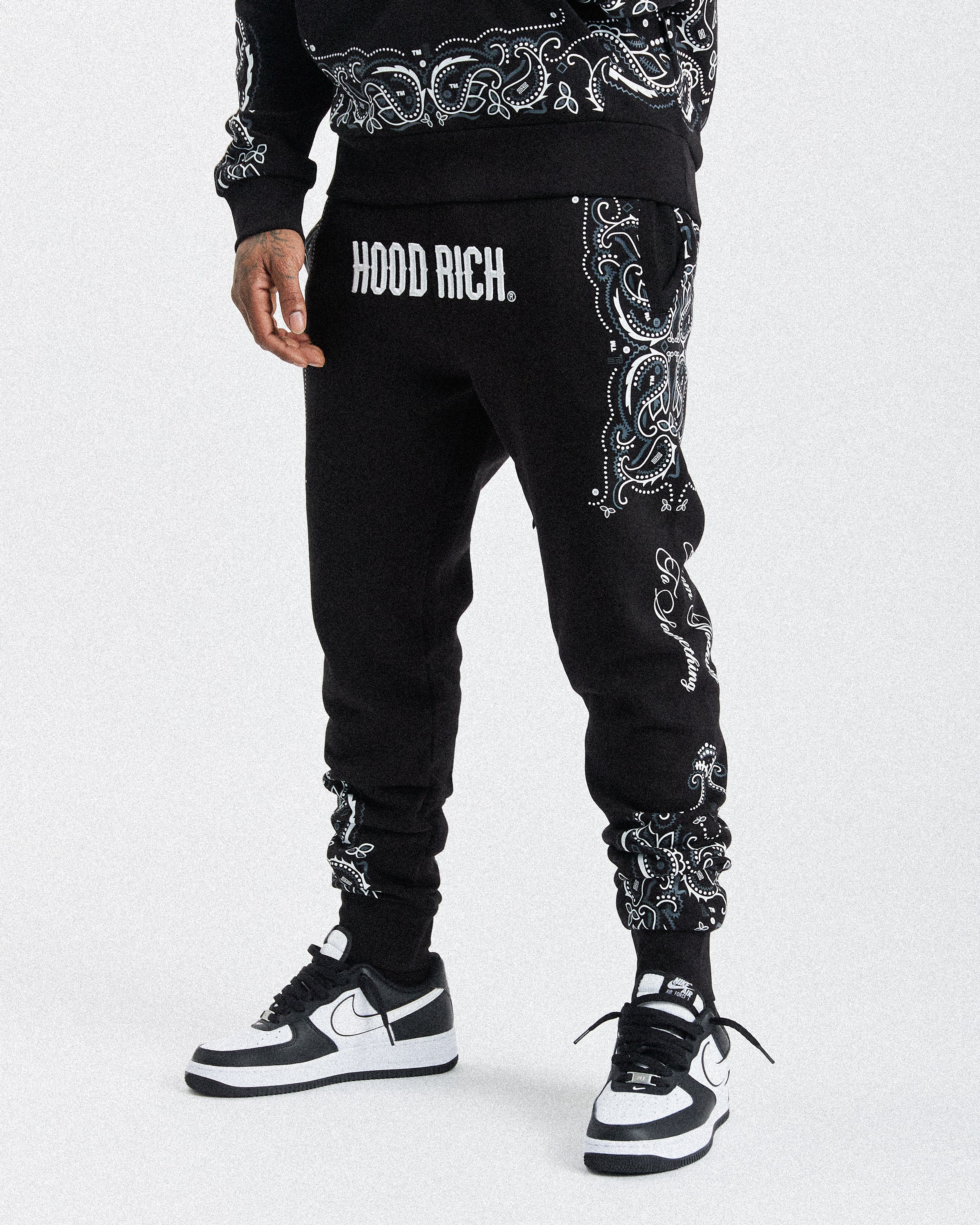 A person wearing black sweatpants with white paisley embroidery down the sides and the text 'HOODRICH.' on the left hip, paired with black and white sneakers against a light gray studio background.