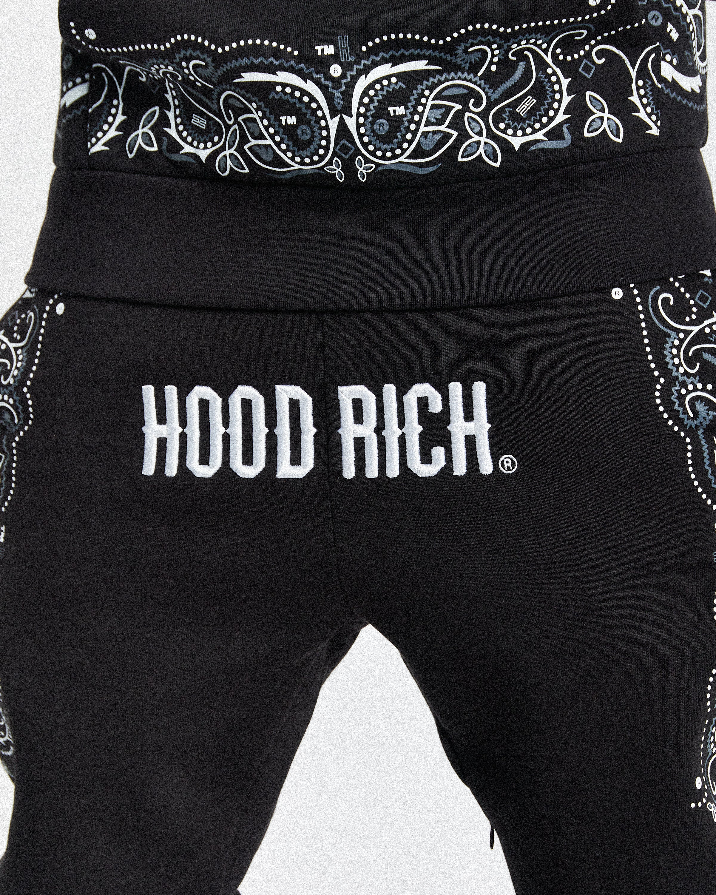Close-up of black sweatpants with a white and gray paisley bandana print around the waistband and side pockets, featuring the word 'HOODRICH.' in bold white letters across the front.