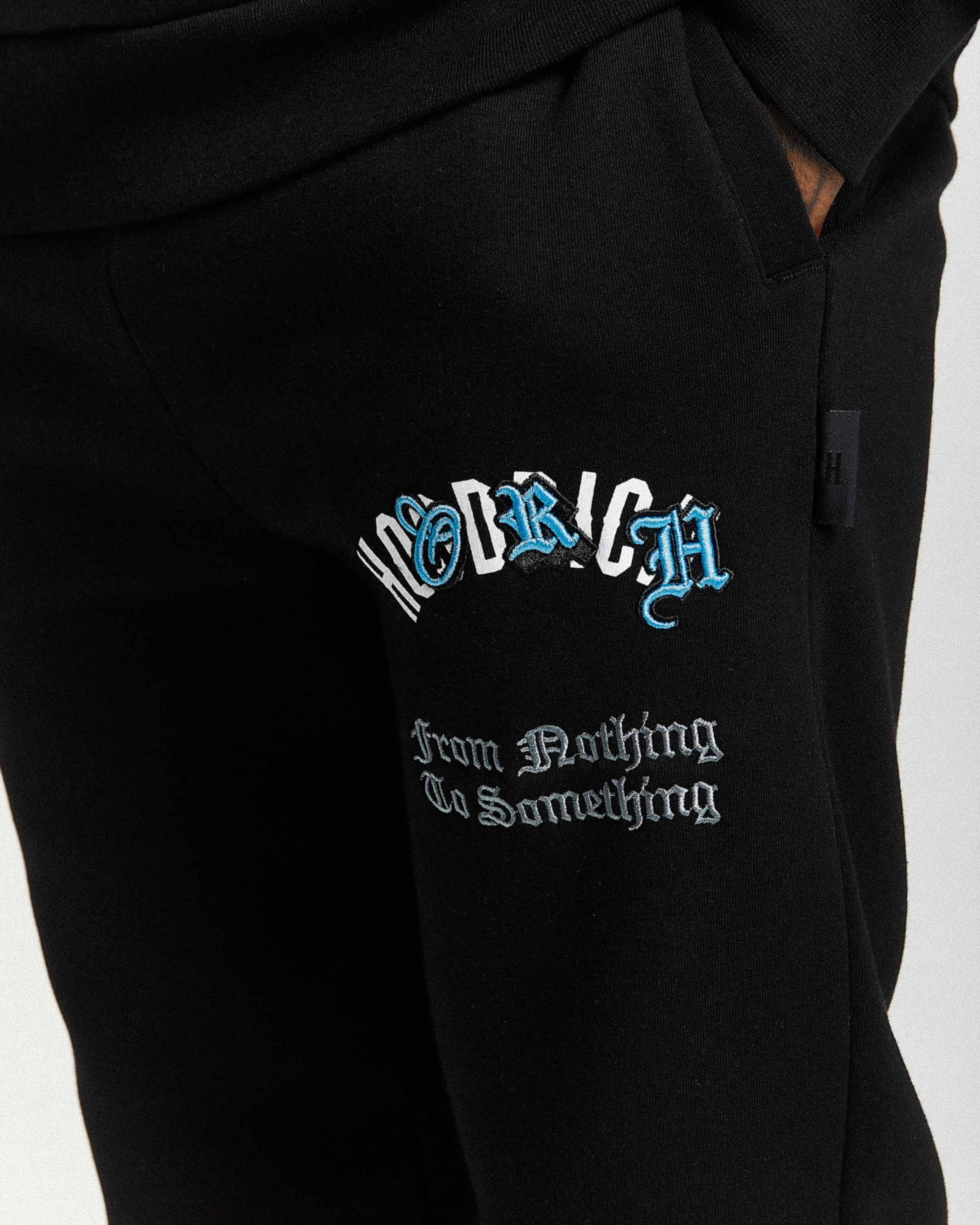 Close-up of black sweatpants with a drawstring waist, featuring a stylized 'Hoodrich' logo in white and blue gothic font and the phrase 'From Nothing To Something' in white gothic text on the left thigh.