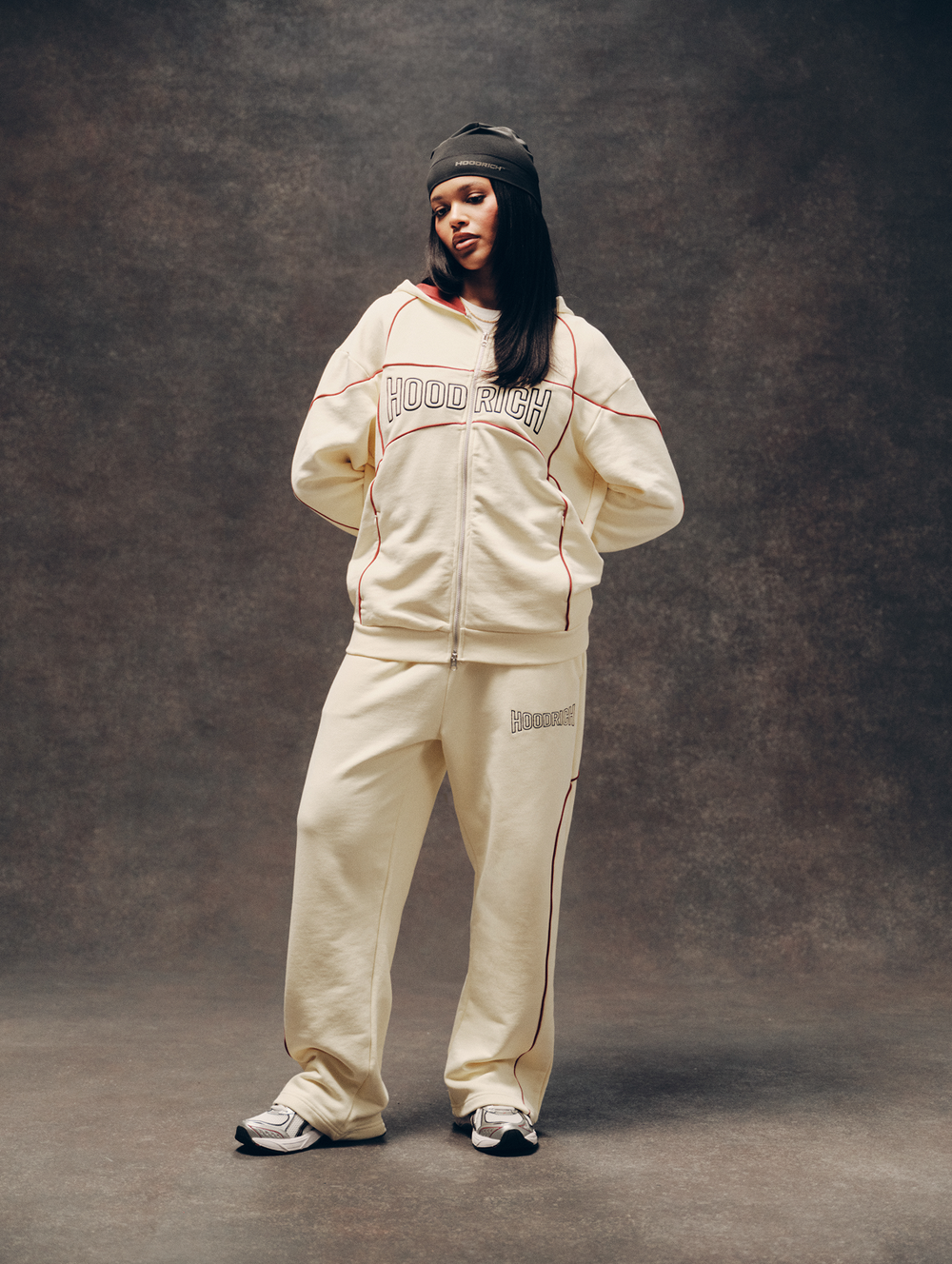 A model wearing a cream-colored 'HOODRICH' zip-up jacket and matching sweatpants with thin red piping, a black beanie, and sneakers, standing against a textured brown studio backdrop.