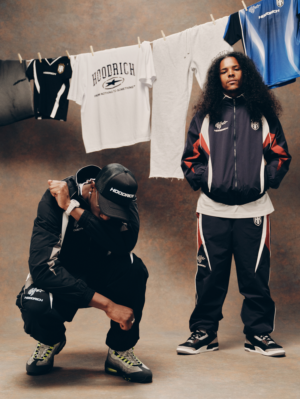 Two models pose in matching 'HOODRICH' streetwear outfits against a textured brown backdrop with vintage sports jerseys hanging on a clothesline. One model is crouching in all black, and the other is standing in a navy, white, and red tracksuit.