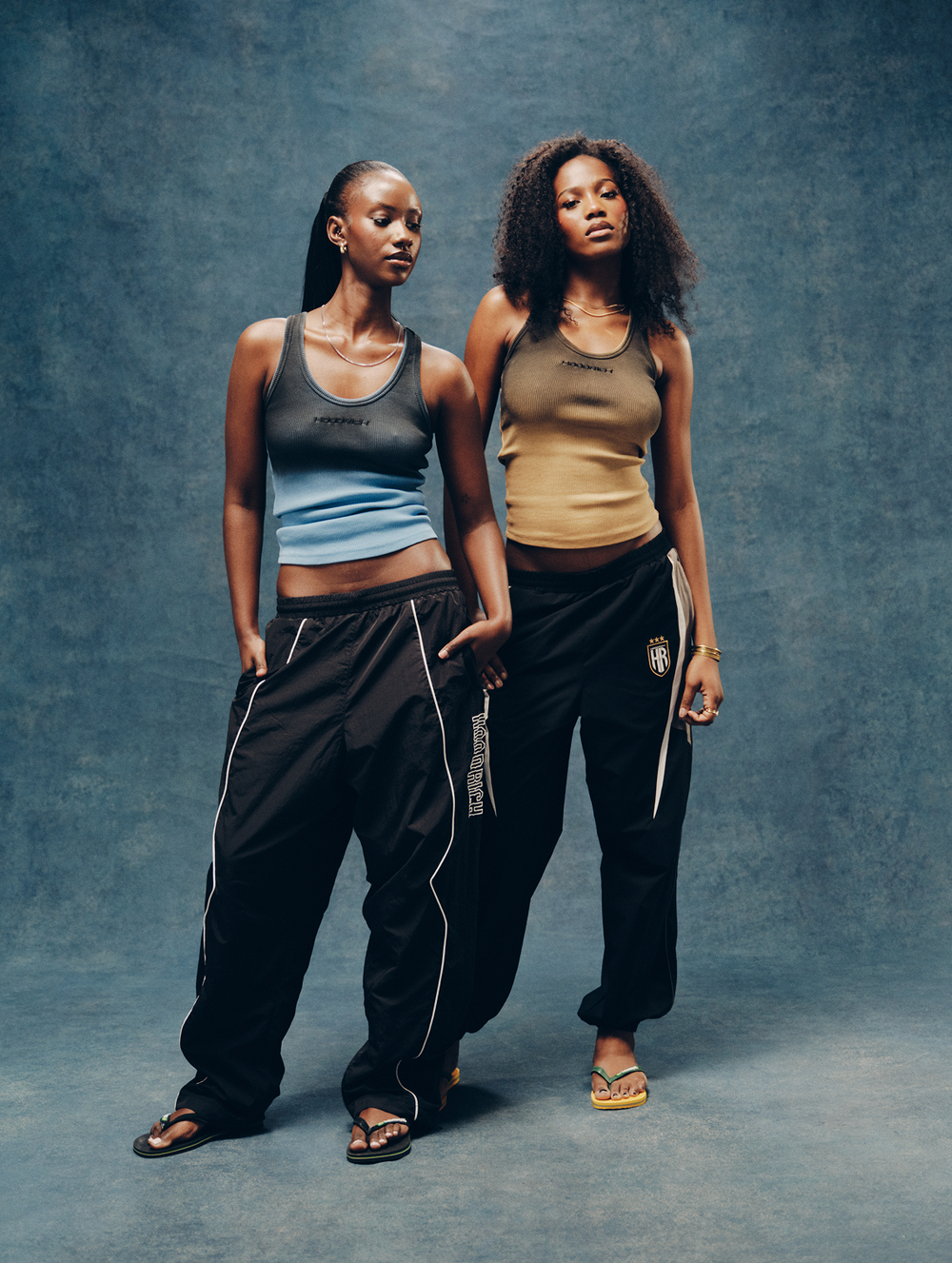 Two models posing in matching streetwear outfits from the brand HOODRICH, featuring black track pants with white piping and tank tops in gray-blue and tan.