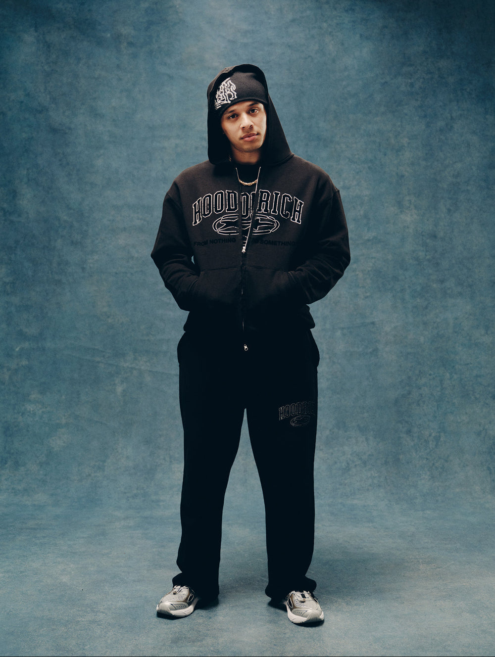 A male model stands against a mottled blue-gray studio backdrop wearing a black hooded zip-up sweatshirt with 'HOODRICH' printed across the chest, matching black sweatpants, a black beanie, and white sneakers.